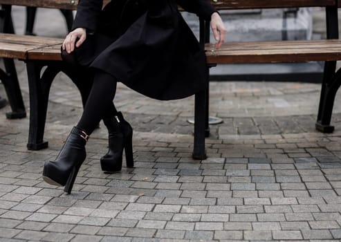 Woman wearing black coat and leather boots and sitting on bench. Trendy casual outfit. Details of everyday look. 