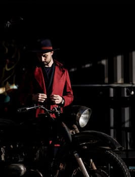 stylish handsome man in a coat and hat near a vintage motorcycle