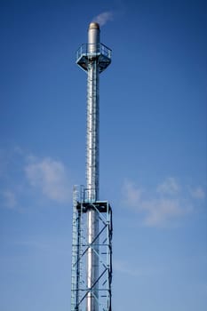 High chimney with smoke coming from. White smoke against the blue sky.