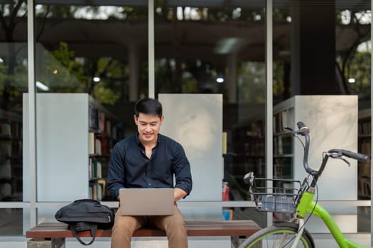 Asian businessman with bicycle using laptop and sitting outside the office building. man commuting on bike go to work. Eco friendly vehicle, sustainable lifestyle concept.