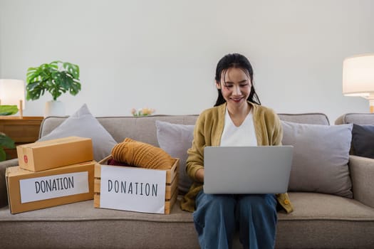 Asian young woman using laptop for donate box with second hand clothes. Donate concept.