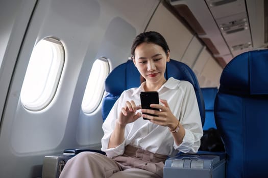 Young Asian woman checks business news on mobile phone, sitting near window in first class on airplane during flight, travel and business concept.