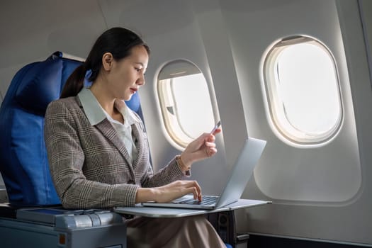 Young Asian woman holding credit card shopping online using website on laptop Sitting near window in first class on airplane during flight, travel and business concept..