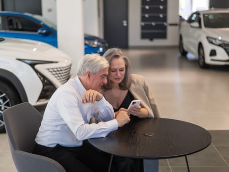 A mature couple is sitting in a car dealership and looking at a smartphone