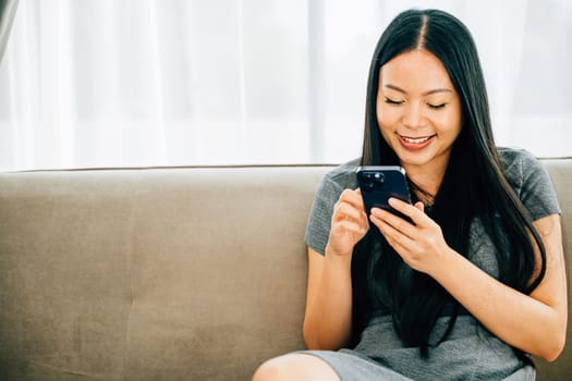 Happy Asian woman sits on a comfortable sofa using her smartphone for texting chatting and online shopping. Enjoying technology for connection and leisure at home.