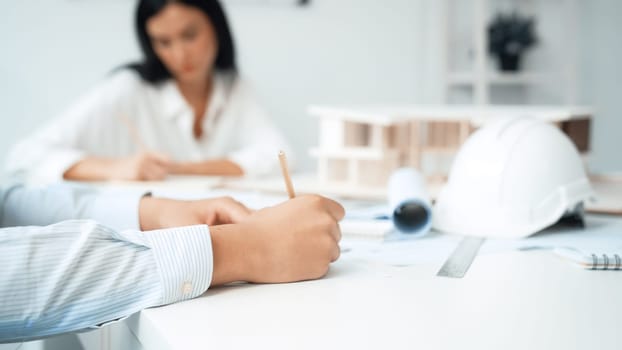 Professional architect engineer drawing blueprint and working together with young beautiful caucasian coworker on meeting table with safety helmet, house model, blueprint scatter around. Immaculate.