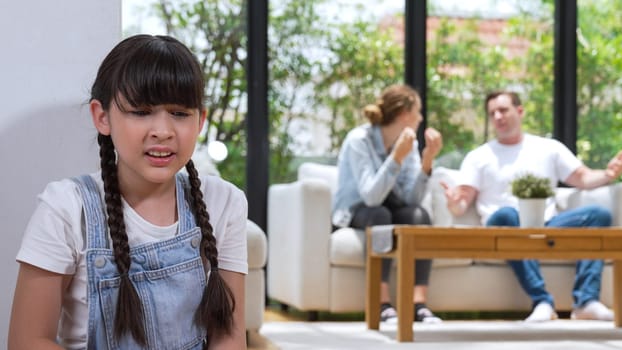 Stressed and unhappy young girl huddle in corner, cover her ears blocking sound of her parent arguing in background. Domestic violence at home and traumatic childhood develop to depression. Synchronos