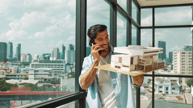 Smart caucasian engineer standing and holding house model while phone calling to manager. Professional manager inspect at architectural model while standing near window. Design concept. Tracery