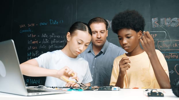 Caucasian teacher talking about electric tool while student fixing robotic model on table with electronic equipment placed. Diverse academic children learning about main board structure. Edification.