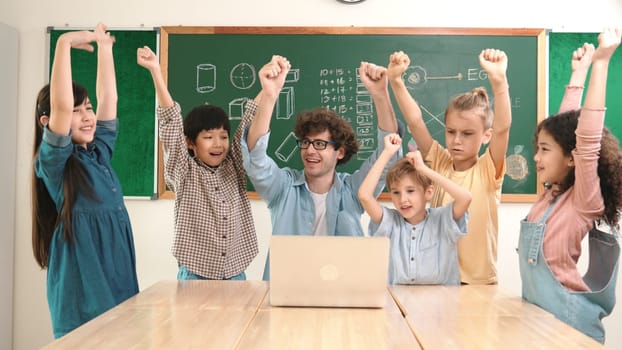 Teacher and multicultural student looking at laptop while celebrate success project and put hands in the air. Group of children smiling to camera while standing at blackboard in classroom. Pedagogy.