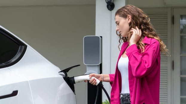 Modern eco-friendly woman recharging electric vehicle from home EV charging station. EV car technology utilized for home resident to future environmental sustainability. Synchronos
