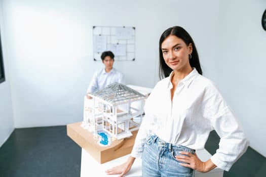 Closeup of female professional architect engineer poses with confident and looks at camera while skilled coworker focus on drawing blueprint. Creative living and design concept. Immaculate.