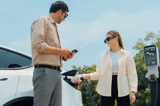 Young couple use smartphone to pay for electricity at public EV car charging station green city park. Modern environmental and sustainable urban lifestyle with EV vehicle. Expedient