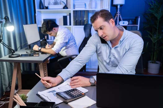 Serious businessman calling on phone with customer to create new project at night time, coworker writing document work sheet on desk. Concept of working on neon blue light modern office. Sellable.