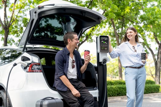 Lovely young couple drinking coffee while recharging battery for electric car during road trip travel EV car in natural forest or national park. Eco friendly travel during vacation and holiday. Exalt