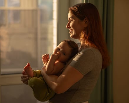 Caucasian woman tenderly holds her newborn son while standing near the window