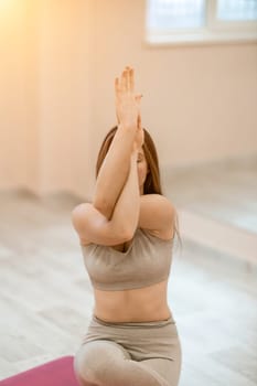 Young woman doing yoga in the gym. A girl with long hair and in a beige tracksuit stands in a cow pose on a pink carpet. A woman performs Gomukhasana