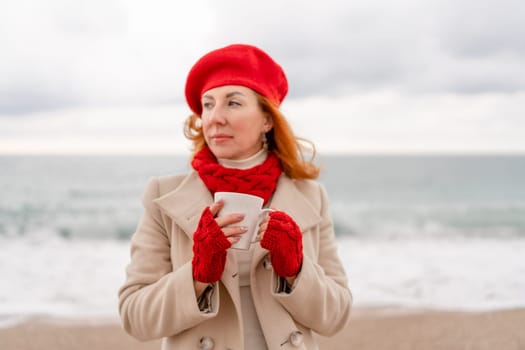 Woman beach in red beret scarf and mitts holding a cup of tea in his hands. Depicting beach relaxation and cozy attire. Walks by the sea.