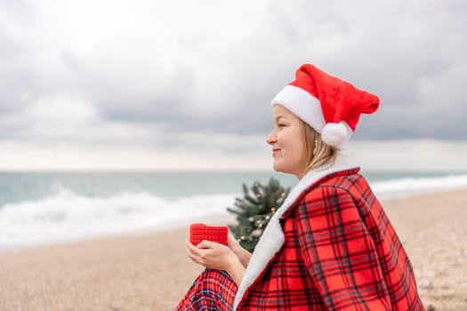 Sea Lady in Santa hat plaid shirt with a red mug in her hands enjoys beach with Christmas tree. Coastal area. Christmas, New Year holidays concep.