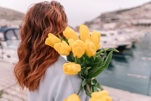 Woman holds yellow tulips in harbor with boats docked in the background., overcast day, yellow sweater, mountains.