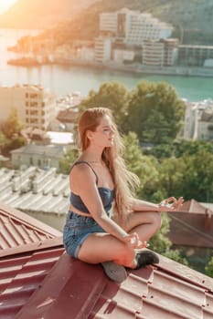 Woman sits on rooftop, enjoys town view and sea mountains. Peaceful rooftop relaxation. Below her, there is a town with several boats visible in the water. Rooftop vantage point