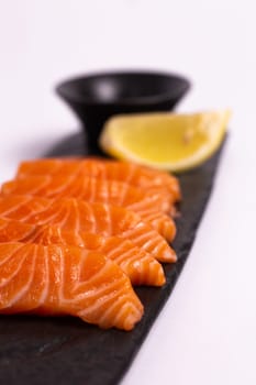 A close-up of a fresh salmon fillet garnished with lemon wedges and soy sauce on a black stone plate. This appetizing seafood dish is isolated on a white background, perfect for a healthy meal.