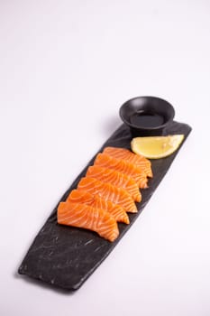 Top view of a delicious and healthy meal of fresh salmon fillet sliced into thin strips and served on a black stone plate with soy sauce and a lemon wedge. The plate is isolated on a white background.