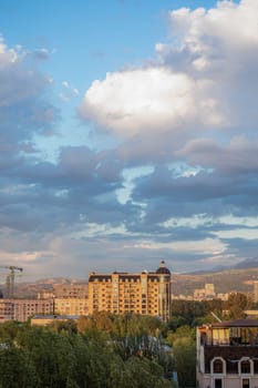 A stunning cityscape with mountains in the backdrop, surrounded by green trees under a blue sky with white clouds.