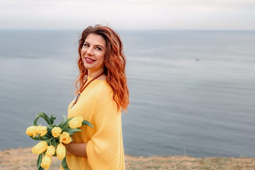 Portrait of a happy woman with long hair against a background of mountains and sea. Holding a bouquet of yellow tulips in her hands, wearing a yellow sweater.