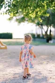 Little girl is walking on the playground under a green tree. Back view. High quality photo