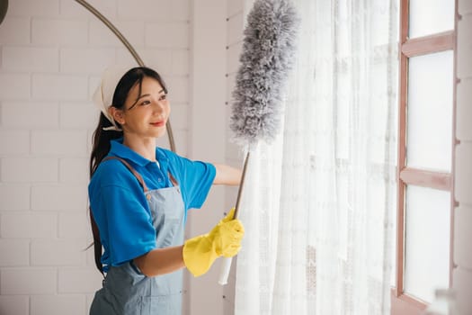 Asian woman's portrait while dusting window blinds. She stands holding duster smiling happily. Occupation involves house hygiene routine cleaning and care. Cleaning service for modern clean household.