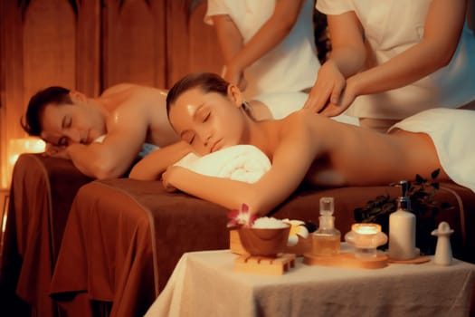 Caucasian couple customer enjoying relaxing anti-stress spa massage and pampering with beauty skin recreation leisure in warm candle lighting ambient salon spa at luxury resort or hotel. Quiescent