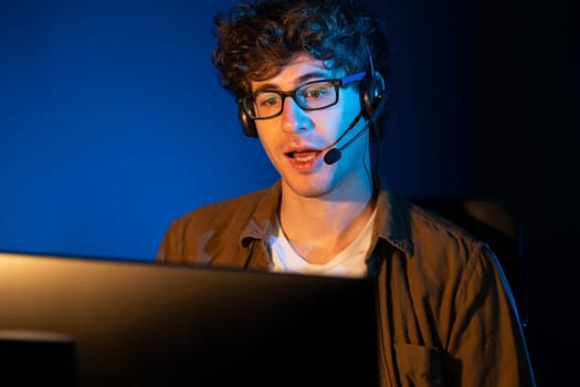 Young creative businessman communicating with listeners on laptop screen or meeting with team member, wearing headphones at neon light modern home office. Concept of working at night time. Gusher.