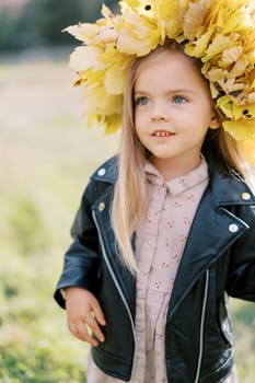 Little girl in a wreath of yellow leaves stands in a field and looks into the distance. High quality photo