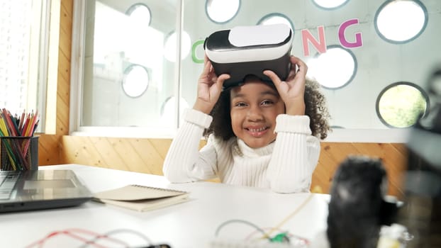 African girl taking off VR glass or head set while looking at camera. Student smiling at camera with colored pencil and laptop placed on table in STEM technology class. Online education. Erudition.