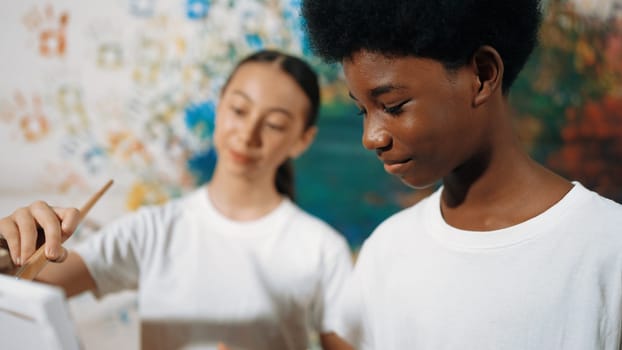 Diverse happy children paint canvas together with colorful stained wall. Smart multicultural student drawing together in art lesson while wearing white shirt. Creative activity concept. Edification.