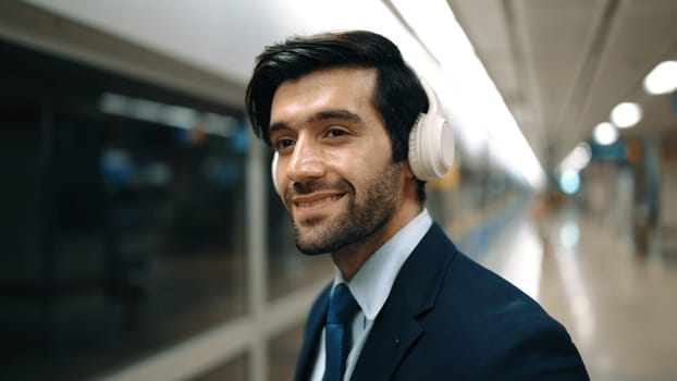 Closeup of smart business man wearing headphone and listening music while standing at train station. Professional executive manager looking at camera while waiting for train or subway. Exultant.
