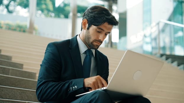 Skilled business man working by using laptop while sitting at stairs. Professional project manager typing and writing marketing plan. Handsome caucasian investor wearing suit with bag. Exultant.