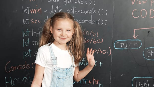 Happy smart caucasian girl smiling at camera while waving hands to camera.while standing at board with engineering prompt or coding, programing system written in STEM technology classroom. Erudition.