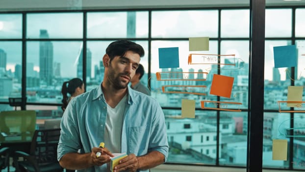 Professional businessman uses sticky notes at glass wall with graph to brainstorming idea while multicultural businesspeople discussing about marketing ideas at office with city view. Tracery