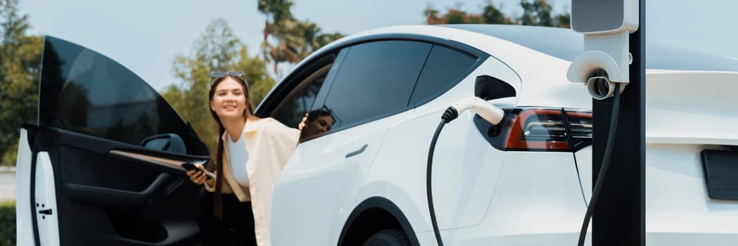 Young woman recharge her EV electric vehicle at green city park parking lot. Urban sustainability lifestyle for environmental friendly EV car with battery charging station. Panorama Expedient