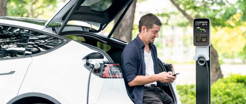 Man using smartphone online banking application to pay for electric car battery charging from EV charging station during vacation holiday road trip at national park or summer forest. Panorama Exalt