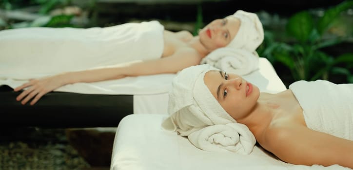 Two beautiful young woman lie on spa bed with white towel while felling in deep relaxation rounded by relaxing and calming nature. Healthy and beauty concept. Blurring background. Tranquility.
