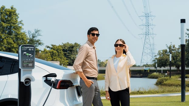 Young couple recharge EV car battery at charging station connected to power grid tower electrical industrial facility as electrical industry for eco friendly vehicle utilization. Expedient