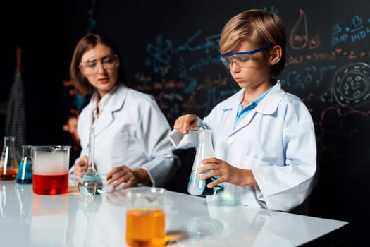 Teacher support schoolboy in laboratory they wear lab coat and glasses stand and experiment about science of chemistry in STEM class. Student tong solid down to yellow liquid in beaker. Erudition.