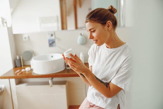 Beautiful young woman taking care of skin by applying moisturizer cream in bathroom