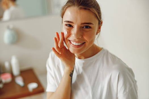 Beautiful young woman taking care of skin by applying moisturizer cream in bathroom