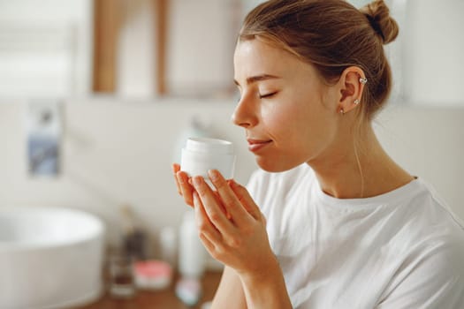 Beautiful young woman taking care of skin by applying moisturizer cream in bathroom