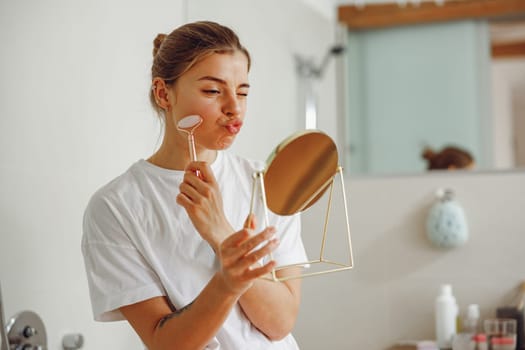 Smiling woman massaging her face with jade roller massager and looking at round cosmetic mirror