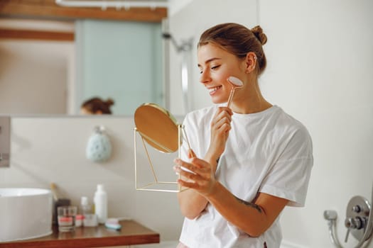 Pretty woman massaging face with jade roller and looking at round cosmetic mirror in bathroom